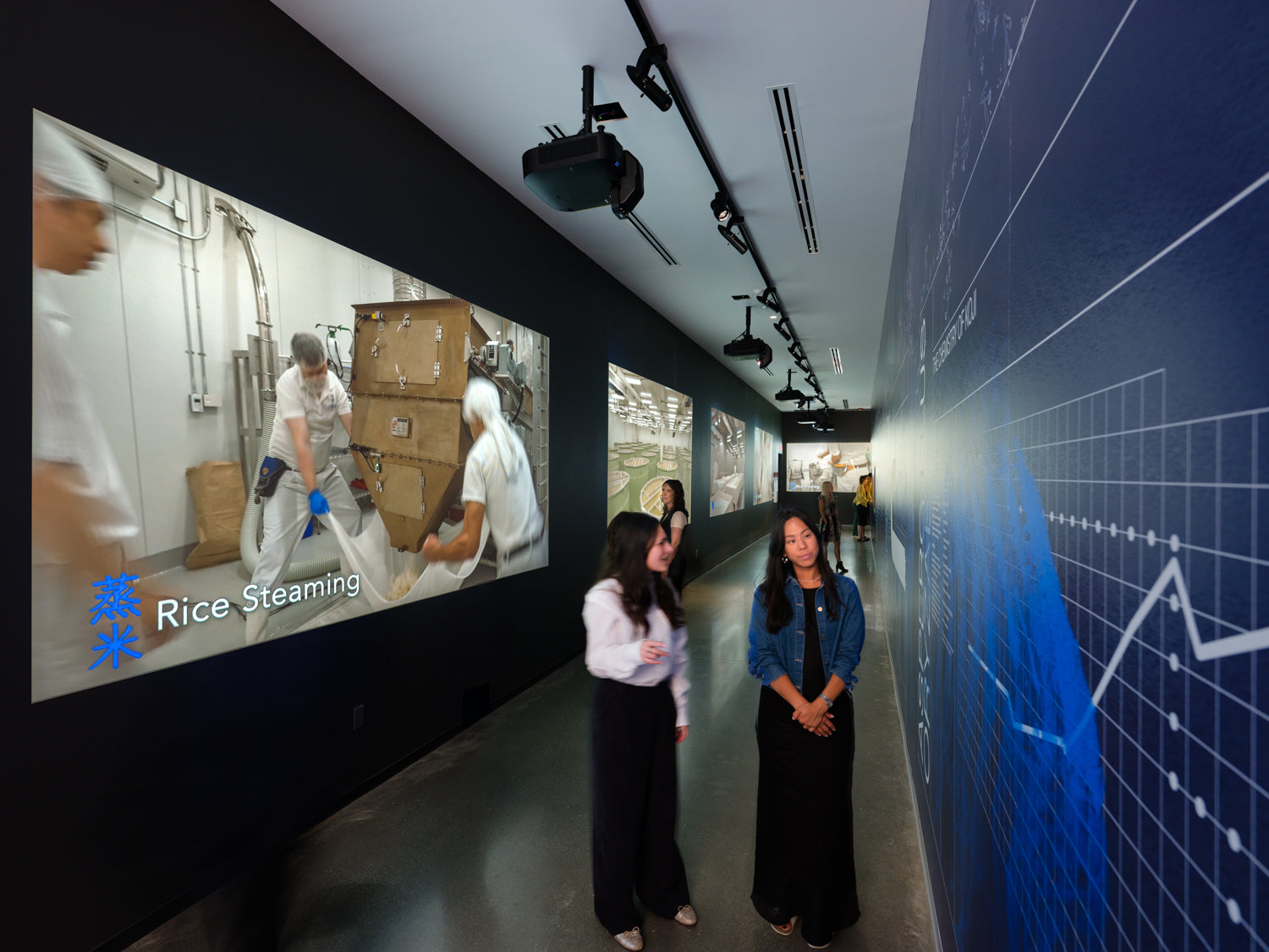 Two women standing in the back corridor with large screens displaying videos, one of which is labeled 'Rice Steaming'.